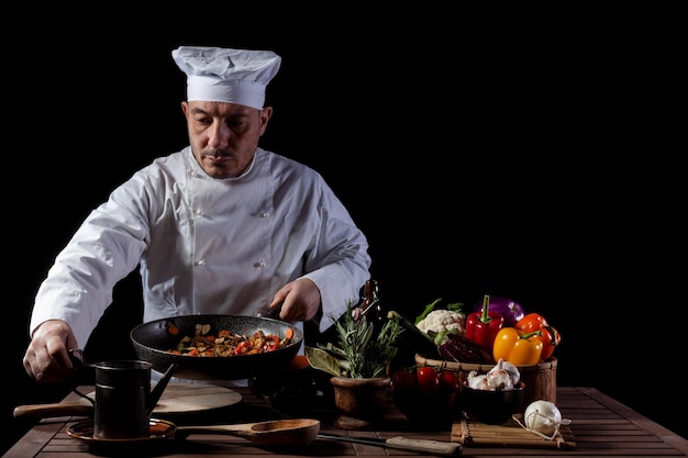Cocinero de sexo masculino con uniforme blanco y sombrero mientras trabajaba en la cocina de un restaurante. Sobre la mesa ingredientes de verduras y hierbas.