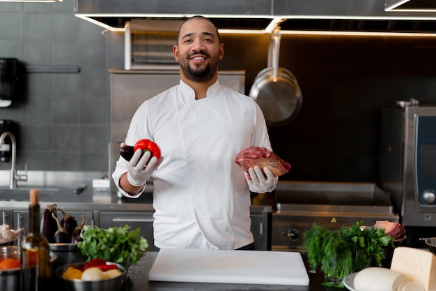 El cocinero sonriente feliz prepara el plato de la carne con las diversas verduras en la cocina. En una mano el hombre sostiene verduras, en la otra un trozo de carne fresca.