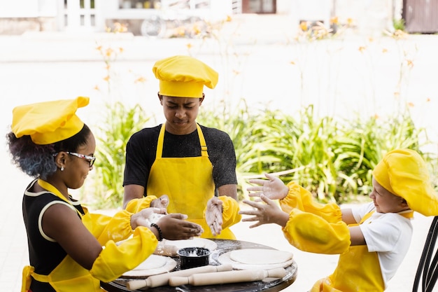 Cocineros multirraciales juegan con harina para hacer masa y se divierten Niños cocineros multinacionales con sombrero de chef y uniforme de delantal amarillo cocinando al aire libre para panadería