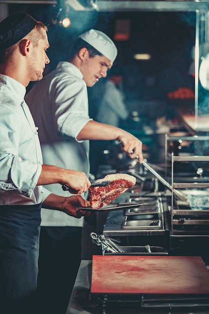 Cocineros ocupados en el trabajo cocinando bistec en el interior de la cocina profesional moderna