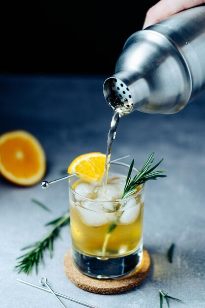 Cocktail de álcool derramando de shaker em vidro com alecrim fatia de laranja e gelo na mesa de madeira