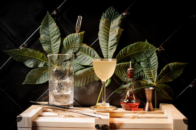 Foto cocktailglas mit gelbem sommercocktail, flasche mit rotem alkohol und bargeräten auf dem hintergrund von grünen blättern