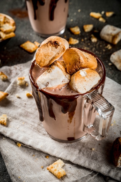 Cocktails tradicionais de outono e inverno, álcool. Cocktail de gemada de chocolate quente na fogueira com biscoitos salgados e marshmallow assado, em duas canecas, na mesa de pedra preta