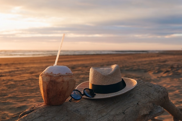 Un coco, gafas de sol y sombrero en el tronco de un árbol en la playa en una hermosa puesta de sol