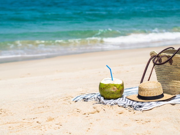 Un coco joven y fresco está listo para comer y una bolsa de paja y un sombrero de paja de mujer en una playa de arena contra un mar azul. Concepto de viajes de vacaciones tropicales. Copia espacio