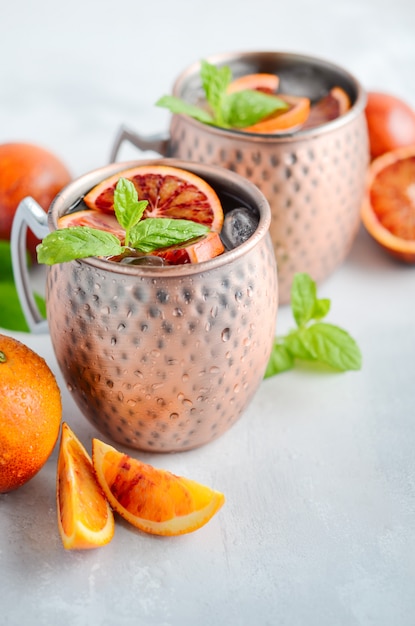 Cóctel del alcohol de la mula de Moscú de la naranja de sangre con las hojas y el hielo de menta fresca en las tazas de cobre en un fondo concreto gris.