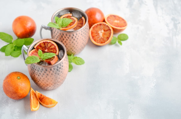 Cóctel del alcohol de la mula de moscú de la naranja de sangre con las hojas y el hielo de menta fresca en las tazas de cobre en un fondo concreto gris.