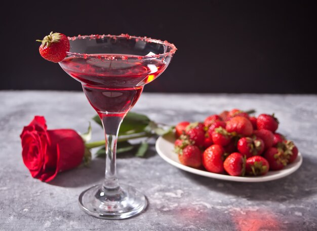Cóctel alcohólico exótico rojo en vidrio transparente, plato con fresas frescas y rosa roja en el fondo de hormigón para una cena romántica.