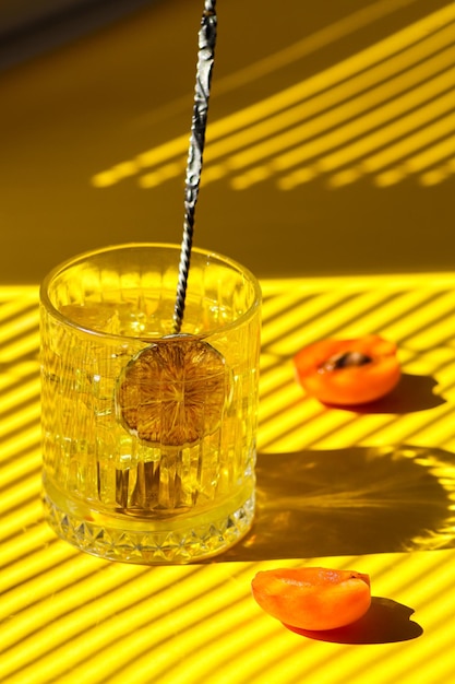 Cóctel amarillo con hielo en un vaso transparente con cuchara de barman, chips de lima y albaricoque fresco.