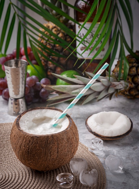 Cóctel coco loco en una mesa de bar con cubitos de hielo, tapa de coco, algunas frutas en el fondo con hojas de palma