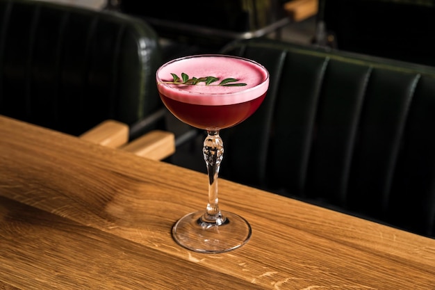 Un cóctel de frambuesa del club de trébol alcohólico agrio rosa con espuma servido en un vaso de cupé adornado con una ramita verde en una mesa de madera en un restaurante o bar foto horizontal