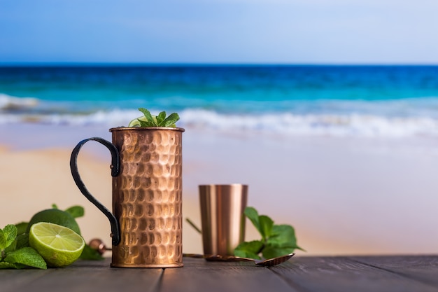 Foto cóctel frío moscow mules con cerveza de jengibre, vodka y lima sobre fondo de playa y mar