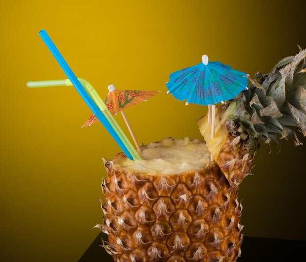 Cóctel de frutas tropicales en piña con pajita y sombrilla. fondo con luz de fondo amarilla