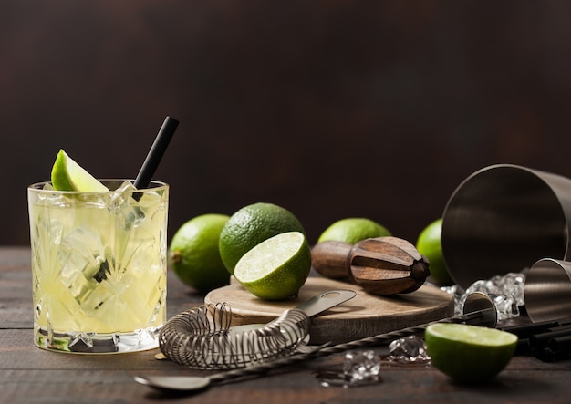 Cóctel Gimlet Kamikaze en copa de cristal con rodaja de limón y hielo sobre superficie de madera con limas frescas y colador con coctelera.