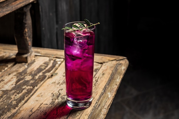 Cóctel de gin tonic hecho con ginebra morada servido con hielo en una copa de cóctel highball adornada con una foto horizontal de una ramita verde