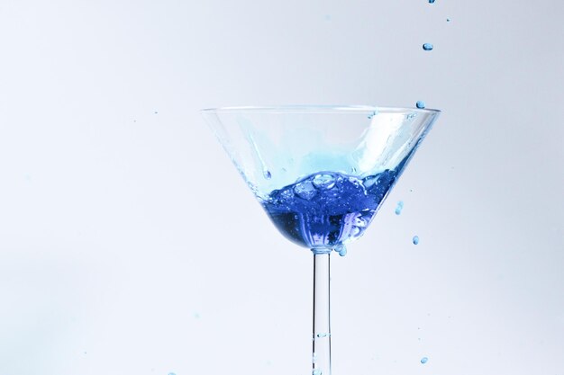 Cóctel con líquido azul en vaso Vaso con agua azul que se vierte con líquido con salpicaduras y gotas Copa de martini llena de alcohol con salpicaduras sobre fondo blanco Concepto de bebida refrescante