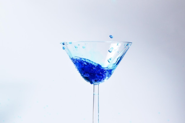 Cóctel con líquido azul en vaso Vaso con agua azul que se vierte con líquido con salpicaduras y gotas Copa de martini llena de alcohol con salpicaduras sobre fondo blanco Concepto de bebida refrescante