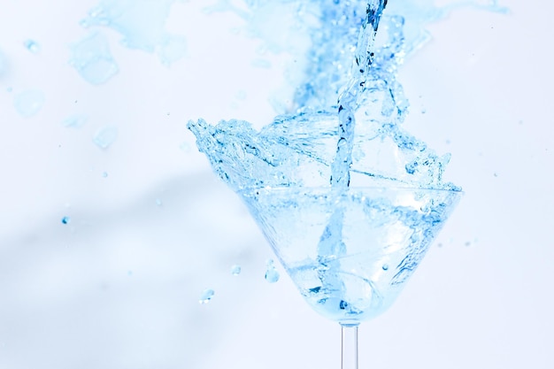 Cóctel con líquido azul en vaso. Vidrio con agua azul vertida con líquido con salpicaduras y gotas. Copa de martini llena de alcohol con salpicaduras sobre fondo blanco. Concepto de bebida refrescante.