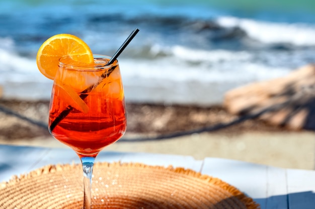 Cóctel rojo con rodaja de naranja y hielo sobre un fondo de mar