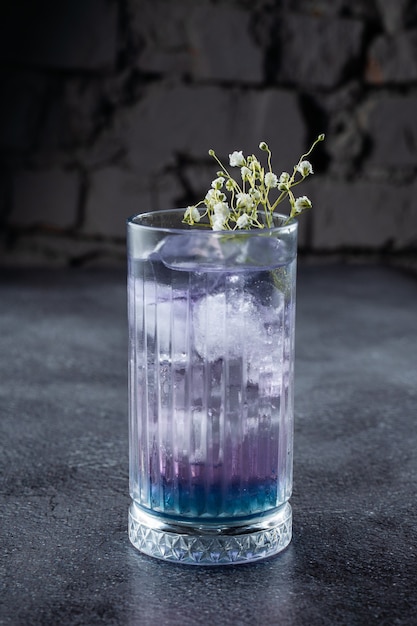 Un cóctel en vaso alto con licor azul y hielo, decorado con flores blancas sobre superficie gris.