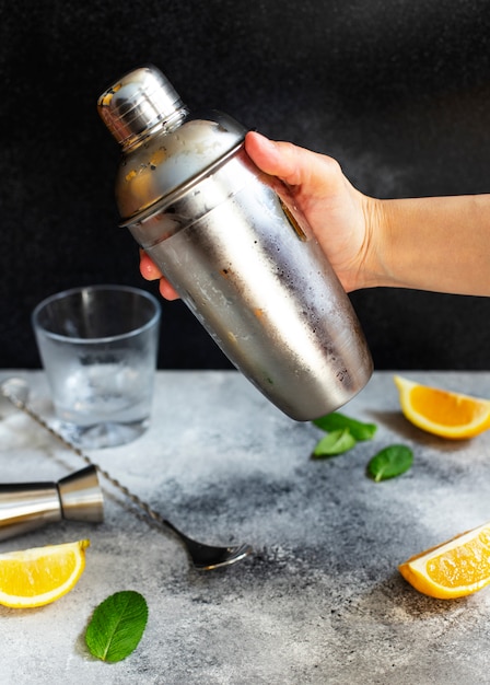 Coctelera y barra de herramientas. Ingredientes para un cóctel alcohólico frío. Coctel Mojito. Limón, menta, hielo, ron. Mano sosteniendo una coctelera. Vista frontal, vertical, fondo gris, espacio de copia