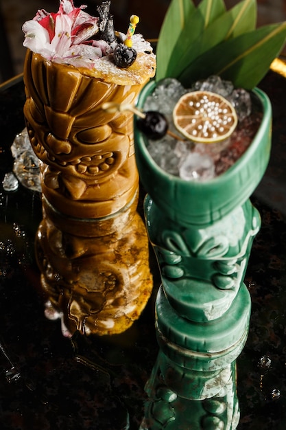 Cócteles de bebida Tiki Dos cócteles en diferentes vasos contra el fondo de un bar decorado con frutas secas y hojas tropicales