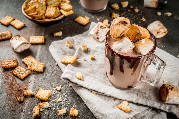 Cócteles tradicionales de otoño e invierno, alcohol. Cóctel de ponche de chocolate caliente con fogatas saladas y malvavisco asado, en dos tazas, sobre una mesa de piedra negra,