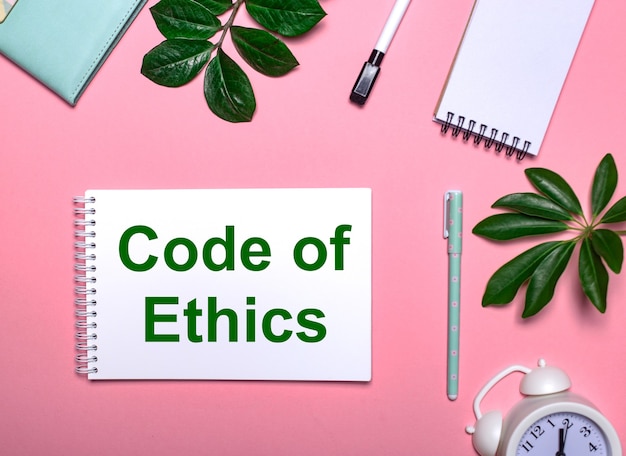 CODE OF ETHICS ist grün auf einem weißen Notizblock auf einem rosa Tisch geschrieben, umgeben von Notizblöcken, Stiften, weißem Wecker und grünen Blättern. Bildungskonzept