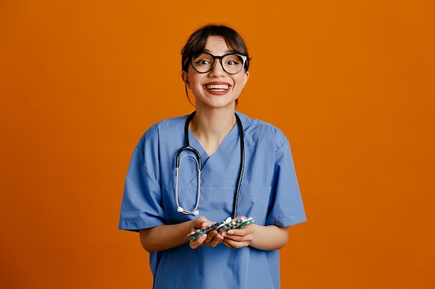 codicioso, tenencia, píldoras, joven, doctora, llevando, uniforme, fith, estetoscopio, aislado, en, naranja, plano de fondo