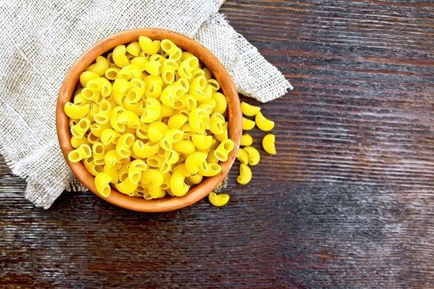 Codo de pasta macarrones en un recipiente de arcilla sobre una servilleta de arpillera en el fondo de la tabla de madera desde arriba