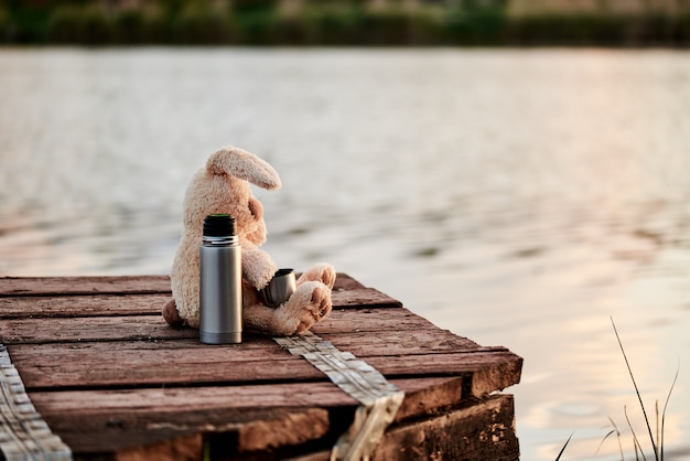 Coelho macio bonito com uma garrafa térmica no cais perto do lago. Copie o espaço.