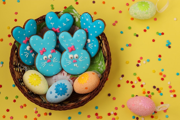 Coelhos azuis engraçados da Páscoa, biscoitos caseiros pintados de gengibre esmaltados em uma cesta de vime sobre uma superfície amarela, vista superior