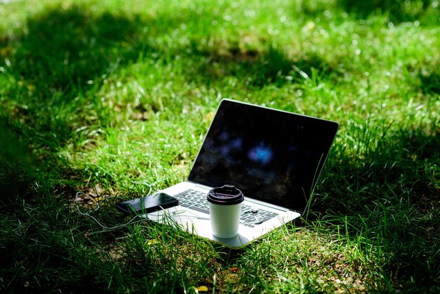 Coffee break al aire libre. Parque de verano. Trabaja y relájate en un entorno natural. Es la hora del café. Café para llevar. Smartphone moderno portátil con auriculares y taza de café sobre la hierba verde. Oficina verde.