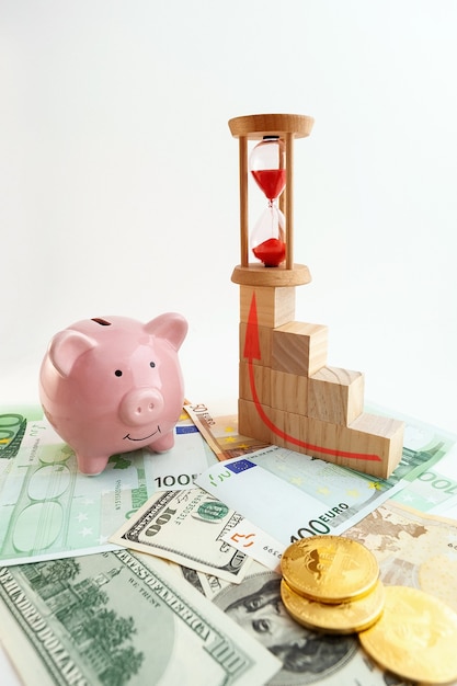 Cofrinho em forma de porco, ampulheta, moedas de bitcoin, cubos de madeira de gráfico crescente, notas de euro e dólares americanos em fundo branco isolado