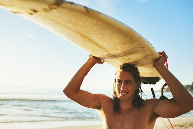 Cogiendo una ola a la vez Captura recortada de un apuesto joven sonriendo mientras lleva su tabla de surf sobre su cabeza en la playa al atardecer