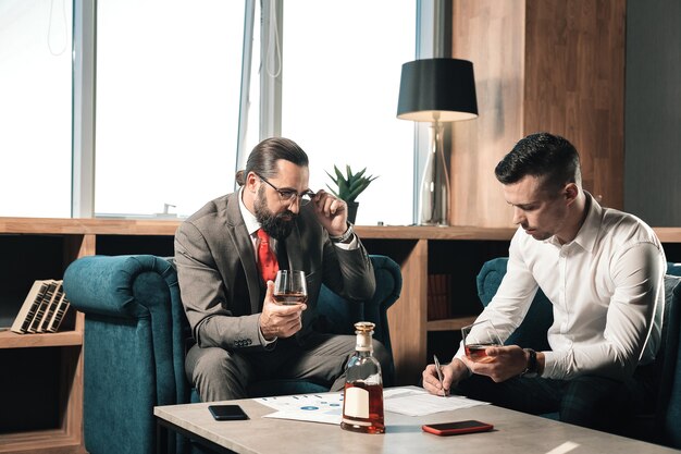 Cognac no escritório. Dois parceiros de negócios inteligentes e bem-sucedidos sentados à mesa no escritório e bebendo conhaque