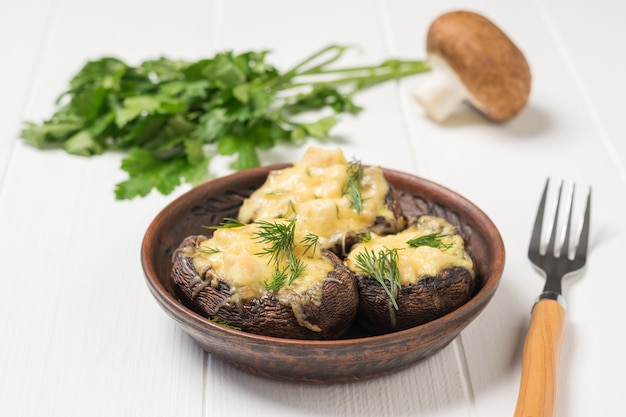 Cogumelos assados em uma tigela de barro e um garfo sobre uma mesa de madeira branca. Um prato de cogumelos assados com queijo.