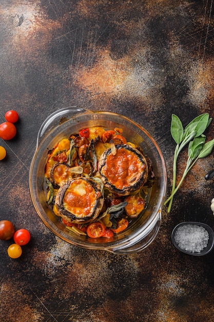 Cogumelos portobello, assados com queijo cheddar, tomate cereja e sálvia em pote de vidro no antigo espaço de vista superior de fundo escuro de metal rústico para texto.