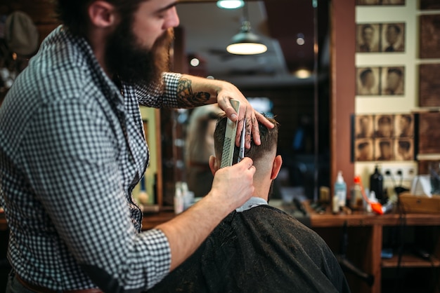 Coiffeur barbudo corte penteado por tesoura. cliente homem com capa preta de salão sentado contra um espelho na barbearia