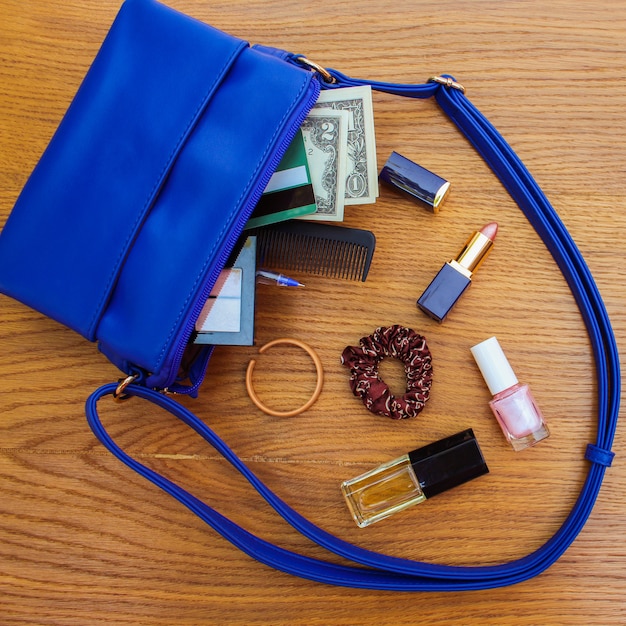 Coisas da bolsa de senhora aberta. bolsa feminina na superfície de madeira. cosméticos e acessórios femininos caíram da bolsa azul.