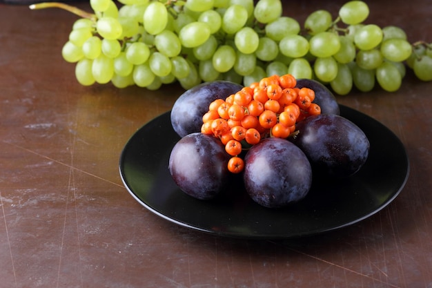 Colección de frutas de otoño crudas y orgánicas del primer plano del jardín Uvas jugosas maduras ciruela y serbal en un plato negro sobre un fondo oscuro