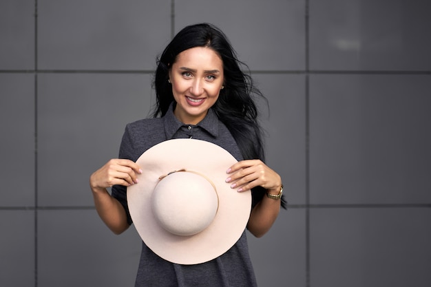 Colección otoño de moda. Feliz chica morena hermosa con elegante vestido gris con sombrero de moda en las manos. Modelo en traje de moda posando en la pared gris. Retrato al aire libre