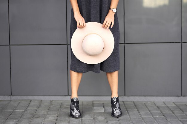 Colección otoño de moda. Feliz chica morena hermosa con elegante vestido gris con sombrero de moda en las manos. Modelo en traje de moda posando sobre fondo de pared gris. Retrato al aire libre