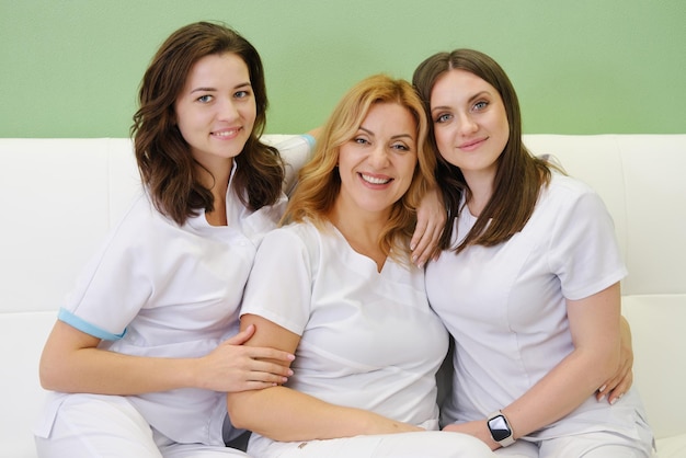 Colegas cosmetólogos con uniformes blancos se sientan juntos en el sofá, sonríen y miran a la cámara El concepto de un equipo de trabajo amigable