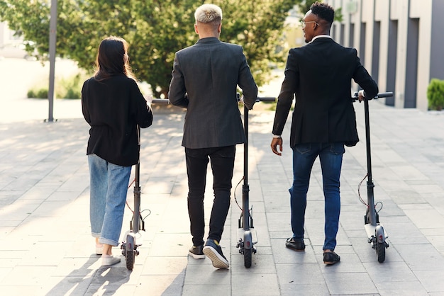 Colegas de escritório multirraciais felizes e confiantes discutindo projeto de negócios vão perto do prédio de escritórios com scooters elétricas