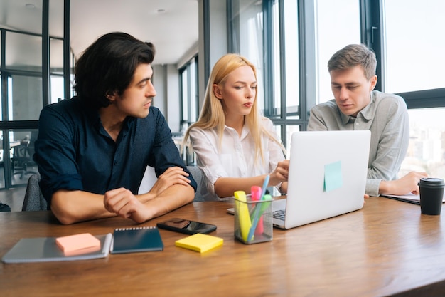 Colegas de negócios de inicialização jovens concentrados, masculinos e femininos, discutindo sentados na mesa, olhando para a tela do laptop na sala de escritório leve pela janela