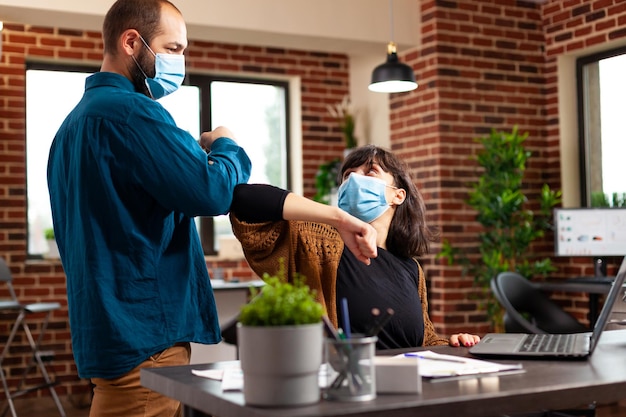 Colegas de trabalho com máscara de proteção médica para evitar infecção com covid19 tocando o cotovelo trabalhando no escritório de inicialização. mulher de negócios digitando estratégia de marketing analisando gráfico de dados