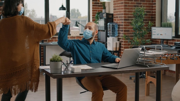 Colegas de trabalho fazendo socos antes de trabalhar no escritório de inicialização. Colegas usando máscaras faciais e trabalhando em projetos de negócios e planejamento estratégico com laptop durante a pandemia de coronavírus.