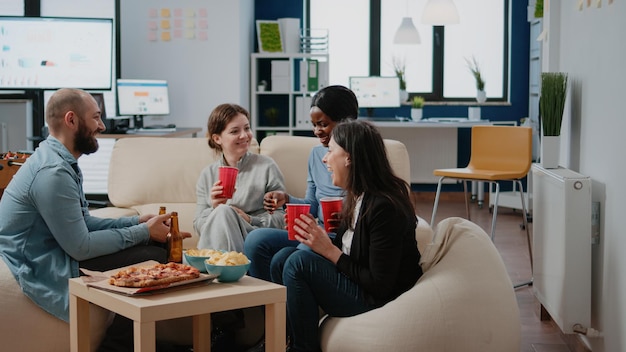 Colegas divirtiéndose con bebidas y bocadillos después del trabajo. Compañeros de trabajo riendo y celebrando la fiesta con botellas de cerveza, bebidas alcohólicas y pizza en la oficina. Compañeros de trabajo disfrutando del ocio