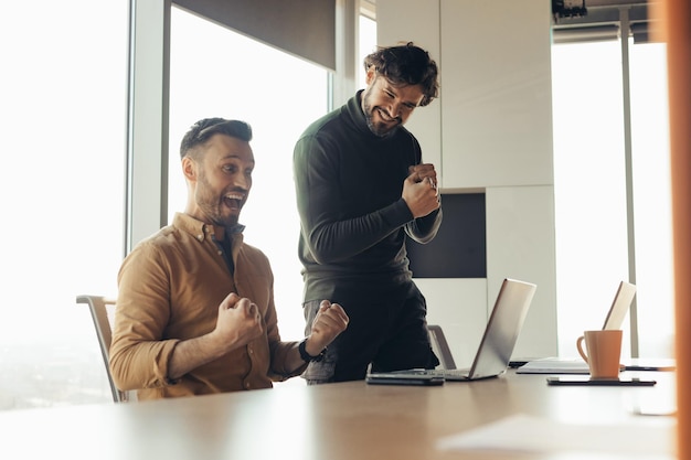 Colegas do sexo masculino animados gesticulando sim comemorando o sucesso nos negócios olhando para a tela do laptop em